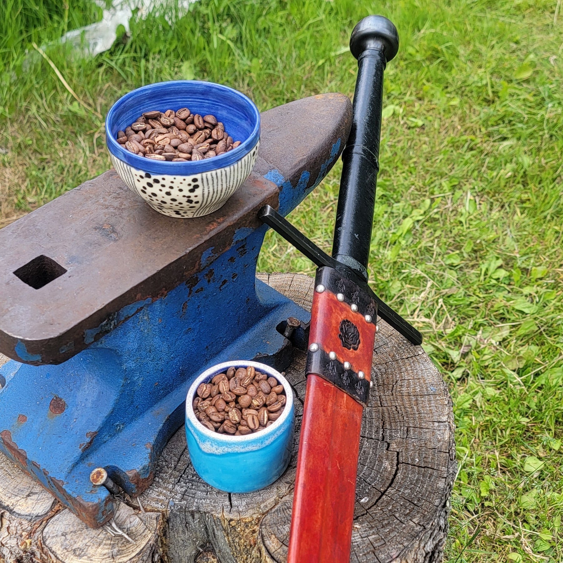 Blue anvil on a stump with two bowls of coffee beans and a red and black sheathed sword outdoors.