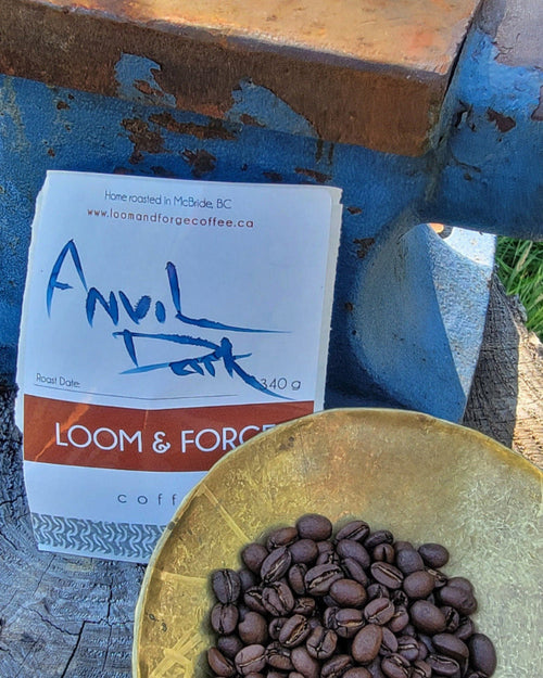 Coffee beans in a bowl with a package labeled 'Anvil Dark' on a rustic metal surface.